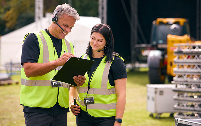 Male And Female Production Team With Headsets Setting Up Outdoor Stage For Music Festival Or Concert